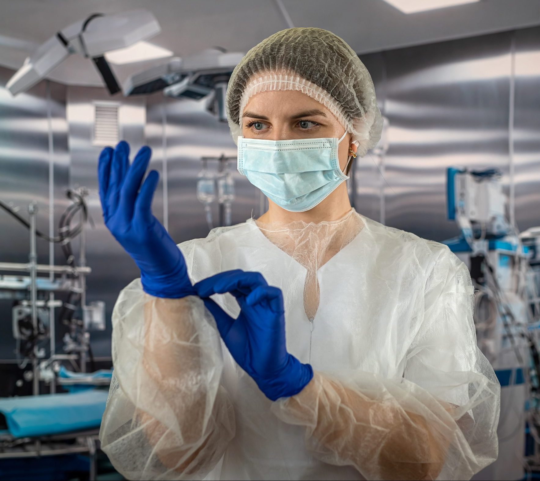 A female nurse wears a blue uniform with a mask while putting on rubber latex blue gloves during a complex surgical operation. medicine. doctors operating room